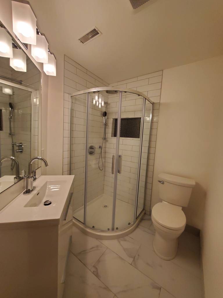 Renovated bathroom with a curved glass corner shower, white wall tile and marble-look floor tile
