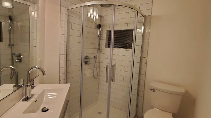 Renovated bathroom with a curved glass corner shower, white wall tile and marble-look floor tile